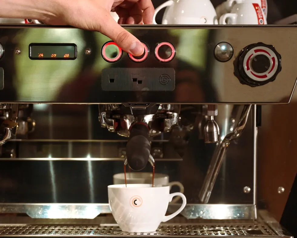Hand pressing a button on a commercial espresso machine while coffee pours into a white Cyso branded cup.